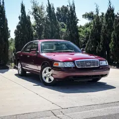 Ciemnoczerwony Ford Crown Victoria, klasyk z USA, zaparkowany na podjeździe wśród wysokich drzew.