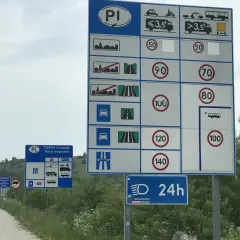 Znaki drogowe informują o ograniczeniu w terenie zabudowanym, opłatach drogowych i limitach prędkości dla różnych pojazdów.