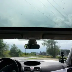 Widok z wnętrza samochodu przez panoramiczny dach. Krople deszczu na szybie, za nią droga i drzewa.