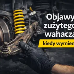 Mechanik w czarnej rękawicy trzyma zużyty wahacz w samochodzie, obok żółtej sprężyny amortyzatora.