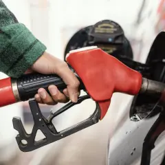 Dłoń w zielonej koszuli trzyma czerwoną dyszę dystrybutora, tankując samochód. Różne rodzaje paliw dostępne na stacji.