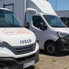 Rząd białych i ciemnych samochodów osobowo dostawczych zaparkowanych na zewnątrz. Na masce białego Iveco widnieje napis "Samochody Używane".