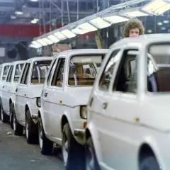 Linia białych samochodów na linii produkcyjnej, z pracownikiem w bordowym płaszczu po lewej.
