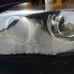 Zaparowany reflektor od środka, z licznymi kropelkami wody na kloszu. Widać odbicie domu i drzew.