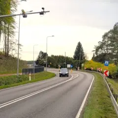 Znak informuje o systemie do pomiaru prędkości w nocy przez policję. Droga kręta, otoczona drzewami.