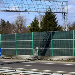 Znak informuje o odcinkowym pomiarze prędkości. Za zieloną barierą dźwiękochłonną widać drzewa i niebo.