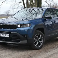 Niebieski SUV klasy premium Jeep Compass na błotnistej drodze, z drzewami w tle.