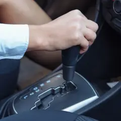 Dłoń na dźwigni **automatycznej skrzyni biegów** w samochodzie. Widoczne oznaczenia biegów: P, R, N, D, 3, 2, L.