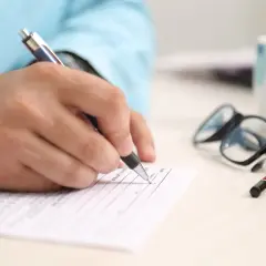 Dłoń pisze na dokumencie, obok okulary i kubek. Zastanawiasz się, co to znaczy RRSO? To roczna rzeczywista stopa oprocentowania.
