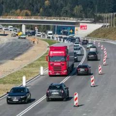 Budowa zakopianki w otoczeniu lasu. Ruch samochodowy, w tym czerwona ciężarówka, odbywa się po nowej drodze z mostem w tle.