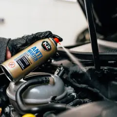 Ręka w rękawiczce rozpyla spray K2 Anti Marten pod maską samochodu, pokazując skuteczne rozwiązanie, czym odstraszyć kuny.