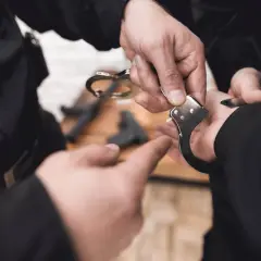 Dłoń policjanta zakłada kajdanki. Czy policjant po służbie może wystawić mandat? Pytanie pozostaje otwarte, gdy widzimy broń na stole.