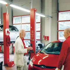Dwóch mężczyzn rozmawia w warsztacie samochodowym. Jeden z nich, ubrany w białą marynarkę, wskazuje na srebrny samochód na podnośniku. Drugi, w czerwonym kombinezonie, stoi przy czerwonym aucie. Widać, że oba pojazdy są **serwisowane w ASO**.
