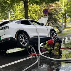 Strażak w akcji, gaszenie samochodu elektrycznego. Woda i węże strażackie wokół białego SUV-a.