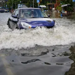 Niebieskie auto walczy z falą wody na zalanej ulicy. W tle widać inne auta po powodzi i autobusy.