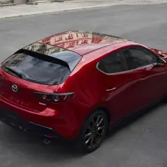 Czerwony Mazda 3 hatchback z czarnymi felgami, odbijający budynki. Elegancki, sportowy design, który odróżnia go od sedana.