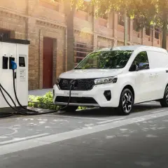 Biały samochód dostawczy podłączony do ładowarki elektrycznej na tle ceglanego budynku.