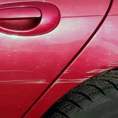 Zarysowanie auta na parkingu. Czerwony lakier z licznymi rysami, widoczna klamka i opona.