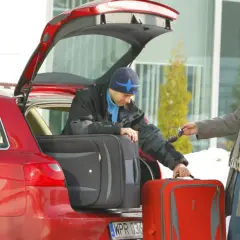 Rodzinne kombi gotowe do wyjazdu. Mężczyźni pakują bagaże, jeden sprawdza coś na kalkulatorze, drugi rozmawia przez telefon.