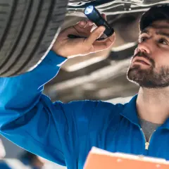 Mechanik w niebieskim kombinezonie oświetla latarką podwozie samochodu, wykonując **serwisowanie samochodu**.