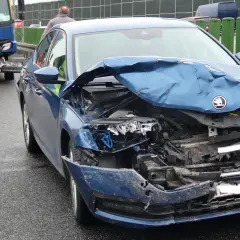 Widok na zniszczony przód niebieskiego samochodu po wypadku samochodowym. Na drodze widać osoby w kamizelkach odblaskowych.