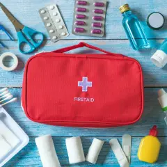 Czerwona torba z napisem FIRSTAID i krzyżem, obok niej wyposażenie apteczki samochodowej: plastry, bandaże, nożyczki, pęseta, leki, płyny.