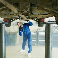 Mechanik w niebieskim kombinezonie bada podwozie samochodu na podnośniku. W powietrzu unosi się lekka mgiełka, przypominająca zapach benzyny w samochodzie.
