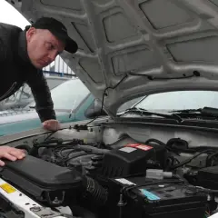 Mężczyzna w czapce z daszkiem pochyla się nad silnikiem białego auta, sprawdzając jego stan. To obrazek z czasów, gdy pierwsze auta były nowością.