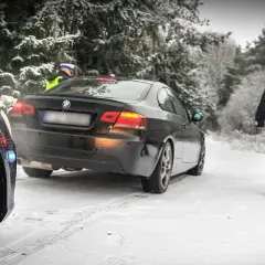 Policjant w kamizelce odblaskowej idzie w stronę czarnego BMW na zaśnieżonej drodze. Kierowca pewnie dostanie mandat za driftowanie.