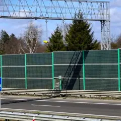Znak informuje o odcinkowym pomiarze prędkości S1. Za zieloną barierą dźwiękochłonną widać drzewa i niebo.