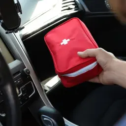 Ręka wkłada czerwoną apteczkę z białym krzyżem do schowka w samochodzie. Czy apteczka jest obowiązkowa? Zawsze warto ją mieć.
