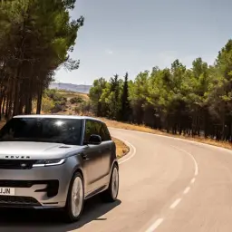 Srebrny Range Rover Sport, jeden z najlepszych SUV-ów, pokonuje zakręt na malowniczej drodze wśród drzew.