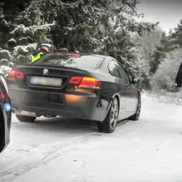 Policjant w kamizelce odblaskowej idzie w stronę czarnego BMW na zaśnieżonej drodze. Kierowca pewnie dostanie mandat za driftowanie.
