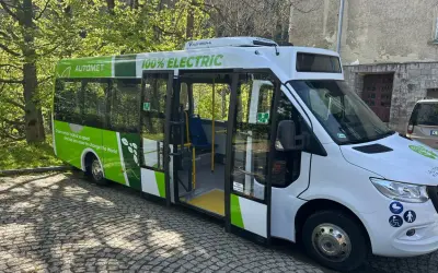 Nowy, zielono-biały bus elektryczny "Automety" z napisem "100% ELECTRIC" stoi na bruku.