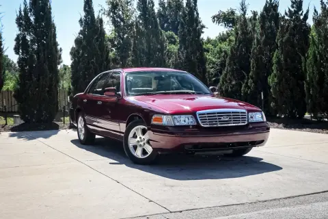 Ciemnoczerwony Ford Crown Victoria, klasyk z USA, zaparkowany na podjeździe wśród wysokich drzew.
