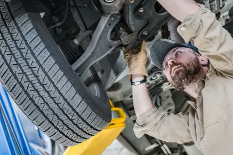 Mechanik w okularach ochronnych wykonuje sprawdzenie zawieszenia na stacji diagnostycznej. Widać jego rękę w rękawicy, która dotyka elementów podwozia.