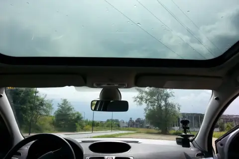 Widok z wnętrza samochodu przez panoramiczny dach. Krople deszczu na szybie, za nią droga i drzewa.