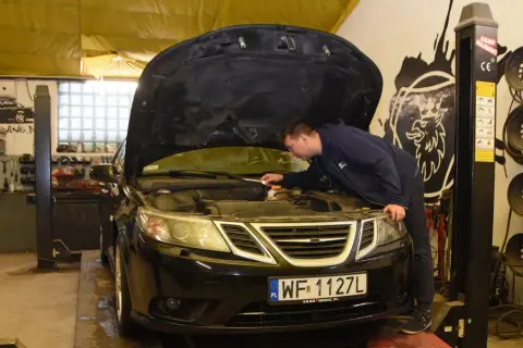 Mechanik sprawdza silnik czarnego Saaba. Na podnośniku warsztatowym, w tle widać narzędzia i części samochodowe.