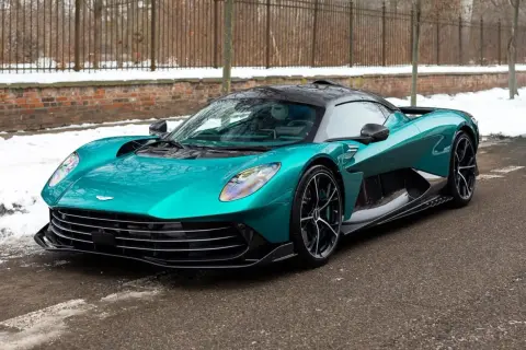 Turkusowy Aston Martin, prawdopodobnie najdroższe auto w Polsce, na tle zimowego krajobrazu z ceglanym murem i płotem.