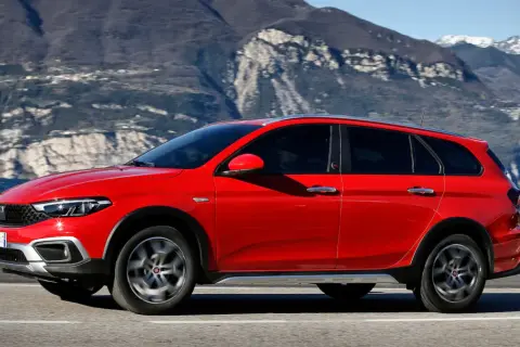 Czerwony Fiat Tipo Cross, wszechstronny samochód z segmentu kombi, przemierza malowniczą drogę nad jeziorem, z górami w tle.