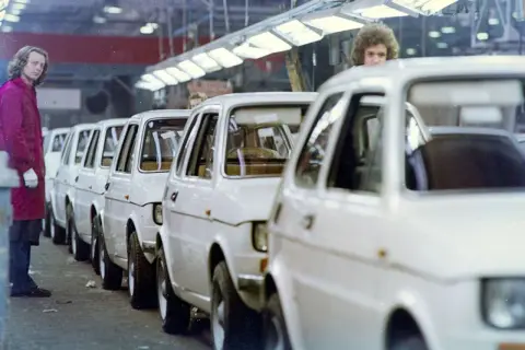 Linia białych samochodów na linii produkcyjnej, z pracownikiem w bordowym płaszczu po lewej.