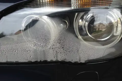 Zaparowany reflektor od środka, z licznymi kropelkami wody na kloszu. Widać odbicie domu i drzew.