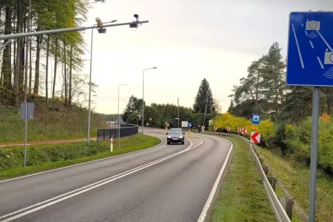 Znak informuje o systemie do pomiaru prędkości w nocy przez policję. Droga kręta, otoczona drzewami.