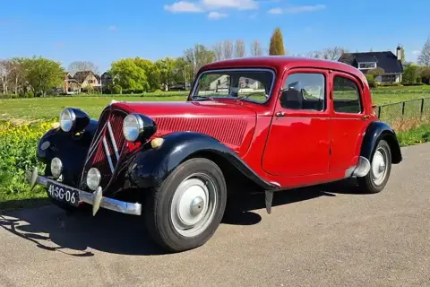 Klasyczny, czerwony Citroen Traction Avant, jeden z kultowych modele stare, zaparkowany na tle zielonej łąki i domów.