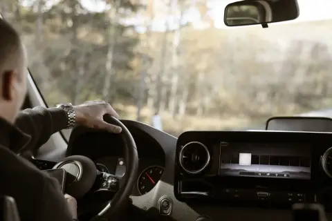 Ręce na kierownicy, zegarek na nadgarstku, a za oknem krajobraz poza miastem. Kierowca cieszy się swobodą i prędkością w terenie niezabudowanym.