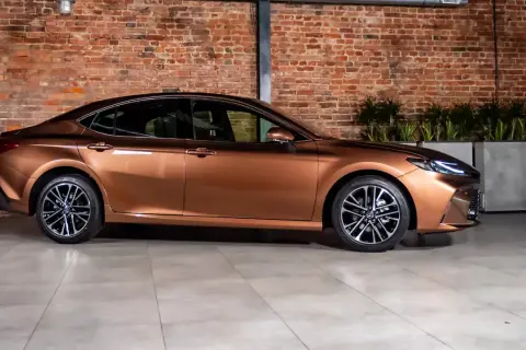 Brązowy sedan o eleganckiej linii, jeden z najładniejszych sedanów, prezentuje się na tle ceglanej ściany i zieleni.