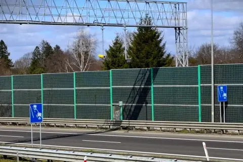 Znak informuje o odcinkowym pomiarze prędkości. Za zieloną barierą dźwiękochłonną widać drzewa i niebo.