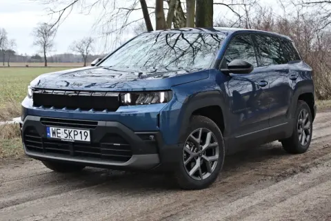 Niebieski SUV klasy premium Jeep Compass na błotnistej drodze, z drzewami w tle.