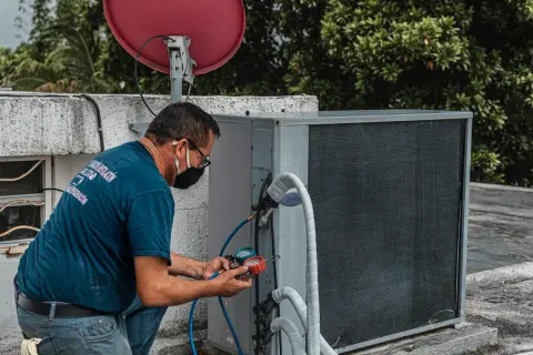 Technik sprawdza klimatyzację, która nie wydaje zimnego powietrza z nawiewu. Na dachu budynku, obok anteny satelitarnej, naprawia urządzenie.