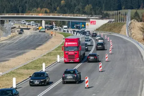 Budowa zakopianki w otoczeniu lasu. Ruch samochodowy, w tym czerwona ciężarówka, odbywa się po nowej drodze z mostem w tle.